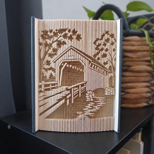 Op de afbeelding: Een gevouwen boekkunstsculptuur met een gedetailleerd driedimensionaal ontwerp van een overdekte brug. De pagina's zijn nauwkeurig gevouwen om de structuur van de brug, omliggende bomen en een stenen pad naar de ingang te vormen.