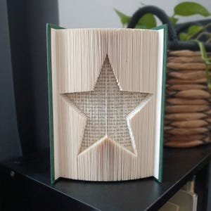 May include: A book with folded pages forming a star shape in the centre. The book has a dark green cover and the pages are cream-coloured with visible black printed text. The pages are precisely folded to create a three-dimensional star silhouette.