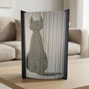 Puede incluir: Una escultura de libro decorativa con la silueta de un gato. La forma del gato se crea con las páginas cortadas de un libro, con un marco negro. El libro se exhibe sobre una superficie de madera.