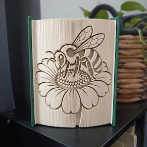 May include: A folded book art sculpture featuring a detailed bee resting on a large flower. The pages are precisely creased to form the image of the insect and the petals. The book has a dark green spine and cover edges, standing upright.