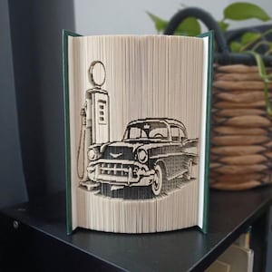 May include: A folded book art sculpture featuring a detailed silhouette of a vintage car parked next to a classic gas pump. The pages are precisely folded to create the three-dimensional image, with the dark green book covers visible on the sides.