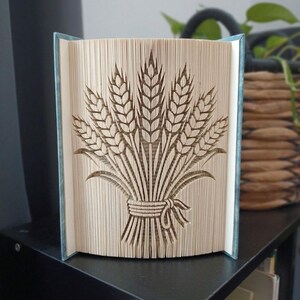 May include: A folded book art sculpture featuring a detailed wheat sheaf design created by precisely folding the individual pages of a hardcover book. The beige paper edges form the shape of multiple stalks of wheat tied with a bow.