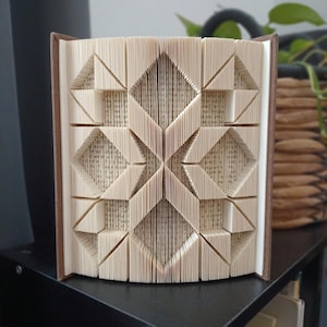 May include: A book with pages folded to create a geometric pattern of diamonds and triangles. The cream-coloured paper edges form a symmetrical, three-dimensional design that stands upright, revealing the printed text on the individual page surfaces.