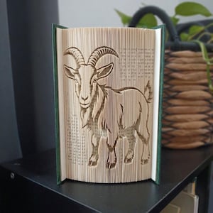 May include: A book sculpture featuring a goat design created by folding the pages of a hardback book. The pages are fanned out to reveal the silhouette of a goat with curved horns, while the dark green book covers remain visible on both sides.