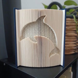 Peut inclure: Une sculpture en livre plié représentant la silhouette d'un dauphin, réalisée en pliant précisément les pages individuelles d'un livre à couverture rigide. Le livre a une couverture bleu foncé et les pages sont crème avec du texte imprimé.