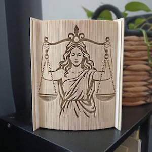 Puede incluir: Una escultura de libro plegado que presenta la figura de la Dama de la Justicia sosteniendo una balanza. Las páginas están dobladas con precisión para crear la silueta de la figura, con el texto del libro visible en los bordes de las páginas.