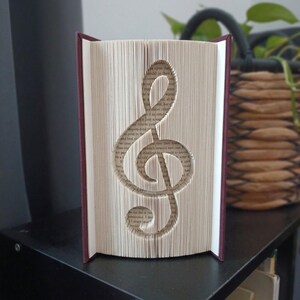 Può includere: Una scultura di libro raffigurante un simbolo di chiave di violino creata piegando con precisione le singole pagine di un libro con copertina rigida. Il libro ha una copertina bordeaux scuro e i bordi delle pagine sono color crema con testo.