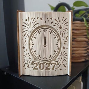Op de afbeelding: Een gevouwen boekkunstsculptuur met een klok die op middernacht staat, omringd door ingewikkelde uitsparingen van vuurwerk, sterren en confetti. De basis toont het jaar 2027 in dikke, uitgesneden cijfers met decoratieve linten.