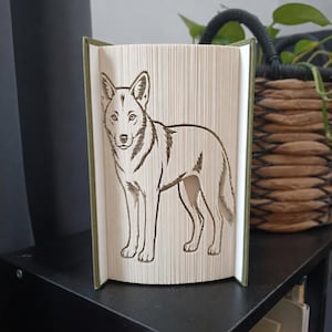 Peut inclure: Une sculpture de livre présentant un design d'art en pages pliées représentant un loup debout. Les pages sont précisément pliées pour créer la silhouette de l'animal, avec les bords sombres du papier formant le contour détaillé du corps et du visage.