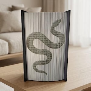 Könnte beinhalten: Eine dekorative Buchskulptur mit einem Schlangenmuster. Die Schlange besteht aus bedruckten Seiten in einem Grau-Weiß-Farbschema. Das Buch hat einen schwarzen Rahmen und steht auf einem Holztisch.