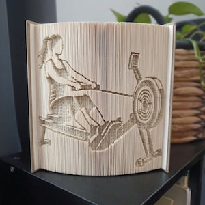 May include: A folded book art sculpture featuring a detailed silhouette of a person using a rowing machine. The pages are precisely folded to create the image of the rower in a seated position with legs extended and arms reaching forward.