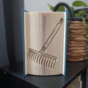 May include: A folded book art sculpture featuring a rake design created by precisely folding the pages of a hardcover book. The book has a teal spine and cover, with the white paper pages forming the distinct shape of a garden rake tool.
