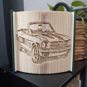 Op de afbeelding: Een boeksculptuur met gevouwen pagina's die de driedimensionale vorm van een vintage cabriolet vormen. De pagina's zijn crèmekleurig met zichtbare tekst en het boek heeft donkergroene covers aan de linker- en rechterkant.