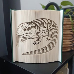 May include: A book sculpture featuring a blue-tongued skink lizard created by precisely folding the pages of a hardback book. The lizard is depicted in profile with its characteristic tongue extended, showcasing detailed scales and striped patterns.