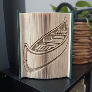 Peut inclure: Une sculpture de livre plié présentant un motif découpé détaillé de canoë. Les pages sont précisément pliées et marquées pour créer la forme tridimensionnelle d'une embarcation avec des sièges intérieurs visibles, encadrée par des couvertures rigides vert foncé.