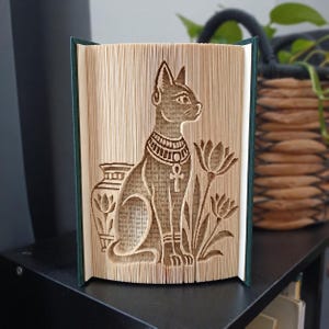 Puede incluir: Una escultura de arte en libro plegado que presenta la silueta detallada de un gato egipcio con un collar con un símbolo ankh. El diseño incluye un jarrón decorativo a la izquierda y dos flores de loto a la derecha, creados doblando con precisión las páginas de un libro de tapa dura.