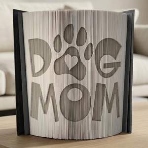 Może przedstawiać: Dekoracyjna rzeźba z książki z napisami "DOG MOM" i odciskiem łapy z sercem wyciętym w stronach. Otwarta książka ukazuje wzór, z czarnym grzbietem. Stoi na drewnianej powierzchni.