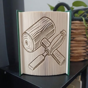 May include: A book art sculpture featuring folded pages that create a three-dimensional image of a wooden log and a chisel. The book has a dark green cover and is displayed in an upright, open position, showcasing the intricate paper folding technique.