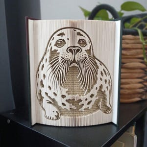 Könnte beinhalten: Eine Buchfaltkunst-Skulptur, die eine detaillierte, dreidimensionale Silhouette eines gefleckten Seehunds zeigt. Die Buchseiten sind präzise gefaltet und geknickt, um das Bild des Seehunds mit Schnurrhaaren, Augen und Flecken zu formen.