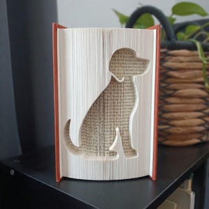 May include: A book sculpture featuring folded pages that form the silhouette of a sitting dog. The book has a reddish-brown cover and white pages with visible black printed text. The dog shape is created by precisely folding the individual pages.