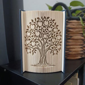 May include: A book sculpture featuring pages folded to create a detailed silhouette of an apple tree. The tree design includes a central trunk, spreading branches, and numerous round apple shapes integrated into the vertical paper edges.