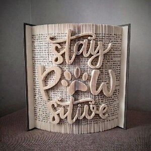 May include: A book sculpture with the words "Stay Paw-sitive" cut into the pages. The text is in a cursive font, with a paw print replacing the "o" in "Paw". The book is open, with the pages fanned out to display the message.