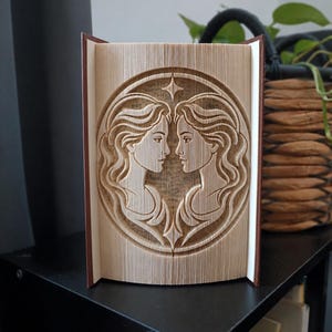 Könnte beinhalten: Eine Buchskulptur aus gefalteten Seiten, die ein dreidimensionales Relief des Sternzeichens Zwillinge bildet. Das Design zeigt zwei gespiegelte weibliche Profile, die sich innerhalb eines kreisförmigen Rahmens gegenüberstehen, gekrönt von einem Stern.