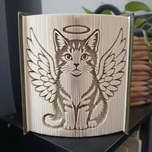 Puede incluir: Una escultura de libro con páginas plegadas que forman la figura de un gato con grandes alas emplumadas y un halo sobre la cabeza. Las páginas son de color crema con texto impreso visible y el libro tiene cubiertas de color verde oscuro.