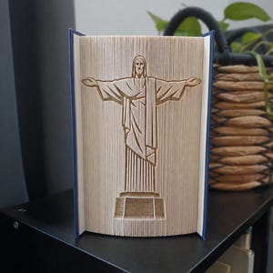 Puede incluir: Una escultura de libro creada mediante el plegado de las páginas de un libro de tapa dura para formar la silueta tridimensional de la estatua del Cristo Redentor. La figura se alza con los brazos extendidos sobre un pedestal, en papel crema.