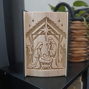 May include: A folded book art sculpture featuring a nativity scene with figures of two adults kneeling beside a manger containing an infant. The design includes a large star above the structure, a cow on the left, and a donkey on the right side.