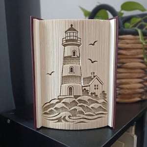 May include: A book sculpture featuring folded pages that form the shape of a lighthouse standing on rocky terrain with waves at the base and birds flying in the sky. The book has a dark purple cover and the pages show visible printed text.