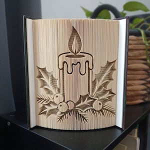 May include: A folded book art sculpture featuring a central candle with a flame, surrounded by holly leaves and berries. The pages are precisely creased to create a three-dimensional relief of the candle and foliage design in beige paper.