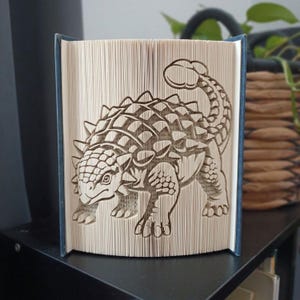Puede incluir: Una escultura de libro plegado con un diseño detallado de un dinosaurio Ankylosaurus. Las páginas del libro están dobladas con precisión para crear la forma tridimensional del dinosaurio acorazado, mostrando su espalda con púas y su cola en forma de maza.