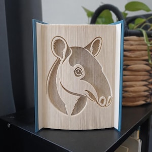 Puede incluir: Una escultura de arte en libro plegado que presenta el perfil detallado de un tapir. Las páginas están dobladas con precisión para formar la cabeza, las orejas y el hocico del animal, creando una imagen tridimensional en los bordes del papel.