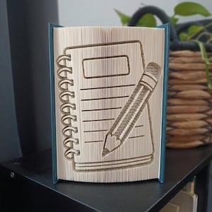 Könnte beinhalten: Eine Buchskulptur aus gefalteten Seiten, die die Form eines Spiralblocks mit einem diagonal aufliegenden Bleistift darstellt. Das Buch hat dunkel-türkisfarbene Einbände und die Seiten sind cremefarben mit erkennbarem Text bedruckt.