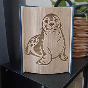 Peut inclure: Une sculpture en livre plié représentant la silhouette détaillée d'un phoque. Les pages sont précisément pliées pour former l'image de l'animal, incluant des moustaches, des yeux et des taches visibles sur le corps. Le livre a une couverture bleue.