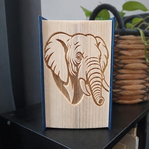Könnte beinhalten: Eine Buchfaltkunst-Skulptur mit einem detaillierten Elefantenkopf-Design, das durch präzises Falten der einzelnen Seiten eines Hardcover-Buches entsteht. Der Elefant ist im Profil mit großen Ohren und einem Rüssel dargestellt.