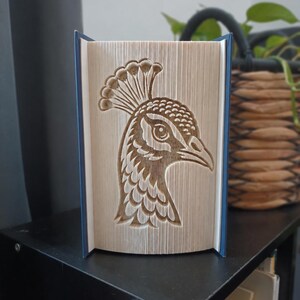 May include: A book sculpture featuring a folded page art design of a peacock head in profile. The pages are precisely creased to form the detailed outline of the bird, including its crest, eye, and beak, set against a dark blue book cover.