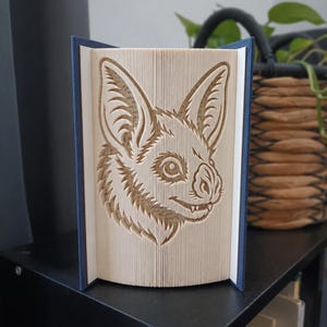 Peut inclure: Une sculpture en livre plié représentant un visage de chauve-souris détaillé, créé en pliant précisément les pages d'un livre à couverture rigide. Le livre possède une couverture bleu foncé et les pages intérieures forment la silhouette d'une tête.