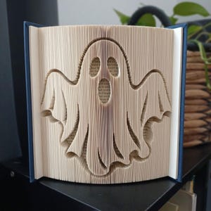 Peut inclure: Une sculpture de livre représentant une forme de fantôme créée en pliant précisément les pages d'un livre à couverture rigide. Les bords des pages couleur crème sont manipulés pour former le contour distinct d'une silhouette spectrale.
