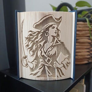 May include: A folded book art sculpture featuring a detailed silhouette of a pirate wearing a tricorn hat and holding a curved sword. The pages are precisely folded to create the image of a figure with long flowing hair and a hoop earring.