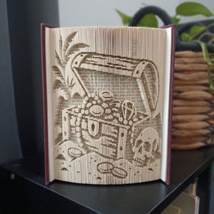 May include: A folded book art sculpture featuring a treasure chest design with an open lid, overflowing with coins, a skull, and palm leaves. The pages are precisely folded to create a three-dimensional relief image of a pirate-themed scene.