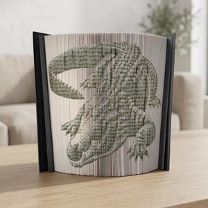 Puede incluir: Una pieza de arte de libro única con un diseño detallado de cocodrilo. La obra de arte se crea doblando las páginas de un libro, mostrando una imagen en escala de grises texturizada del reptil. El libro tiene una columna vertebral negra y se exhibe sobre una superficie de madera.