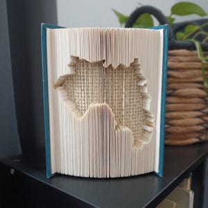 Könnte beinhalten: Eine Buchfaltkunst-Skulptur mit einem türkisfarbenen Einband und weißen Seiten. Die Seiten sind präzise gefaltet, um eine dreidimensionale Silhouette des Kontinents Afrika zu bilden, die von der Vorderseite des offenen Buches aus sichtbar ist.