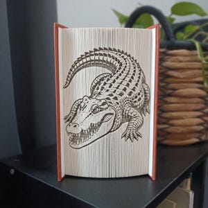 May include: A book sculpture featuring a folded page design that creates a detailed image of an alligator. The pages are precisely creased to form the textured scales, sharp teeth, and curved tail of the reptile, with a reddish-brown spine.