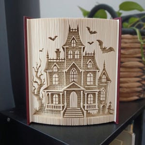 Peut inclure: Une sculpture en livre plié représentant une maison hantée détaillée avec plusieurs niveaux, des fenêtres en arc et un porche. Les pages sont minutieusement pliées pour former la structure, agrémentée de silhouettes de chauves-souris.