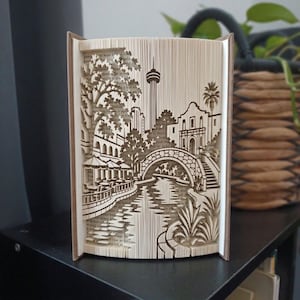Könnte beinhalten: Eine gefaltete Buchkunst-Skulptur mit einem detaillierten Scherenschnitt-Design des San Antonio River Walk, einschließlich des Tower of the Americas, des Alamo, einer Steinbrücke, Palmen und Essbereichen am Flussufer mit Sonnenschirmen.