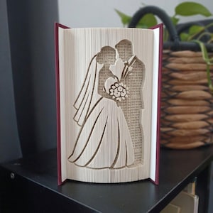 May include: A folded book art sculpture featuring the silhouette of a couple in wedding attire. The pages are precisely folded to create the shape of a person in a gown with a veil and a person in a suit holding a bouquet of flowers.