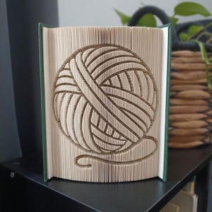 Pode incluir: Uma escultura de livro dobrado com um design de novelo de lã, criado dobrando com precisão as páginas individuais de um livro de capa dura. O livro tem uma capa verde-escura e páginas cor de creme, com o padrão de lã claramente visível.