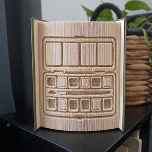 Könnte beinhalten: Eine Buchskulptur mit gefalteten Seiten, die die Form eines geöffneten Aquarellfarbkastens bilden. Das Design umfasst einen rechteckigen Deckel, eine Reihe von vier großen Farbnäpfchen, acht kleine Farbnäpfchen und einen Pinsel.